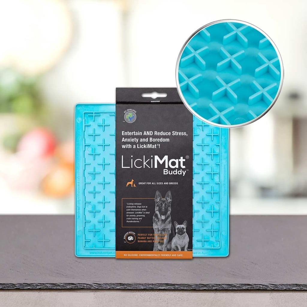 Modra Podloga LickiMat® - BUDDY by PLATINUM je na mizi, na embalaži sta dva psa in informacije o izdelku. Vložek poudarja njeno križno površino za počasno hranjenje psa. Ozadje je zamegljeno.