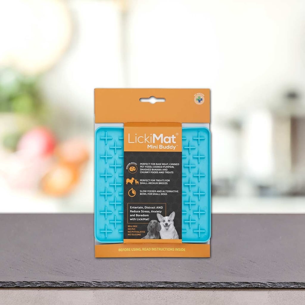 Podloga LickiMat® - BUDDY by PLATINUM v modri barvi z oranžno embalažo stoji pokonci na pultu, idealna za poticanje sporog hranjenja psa, na zamegljenem kuhinjskem ozadju.