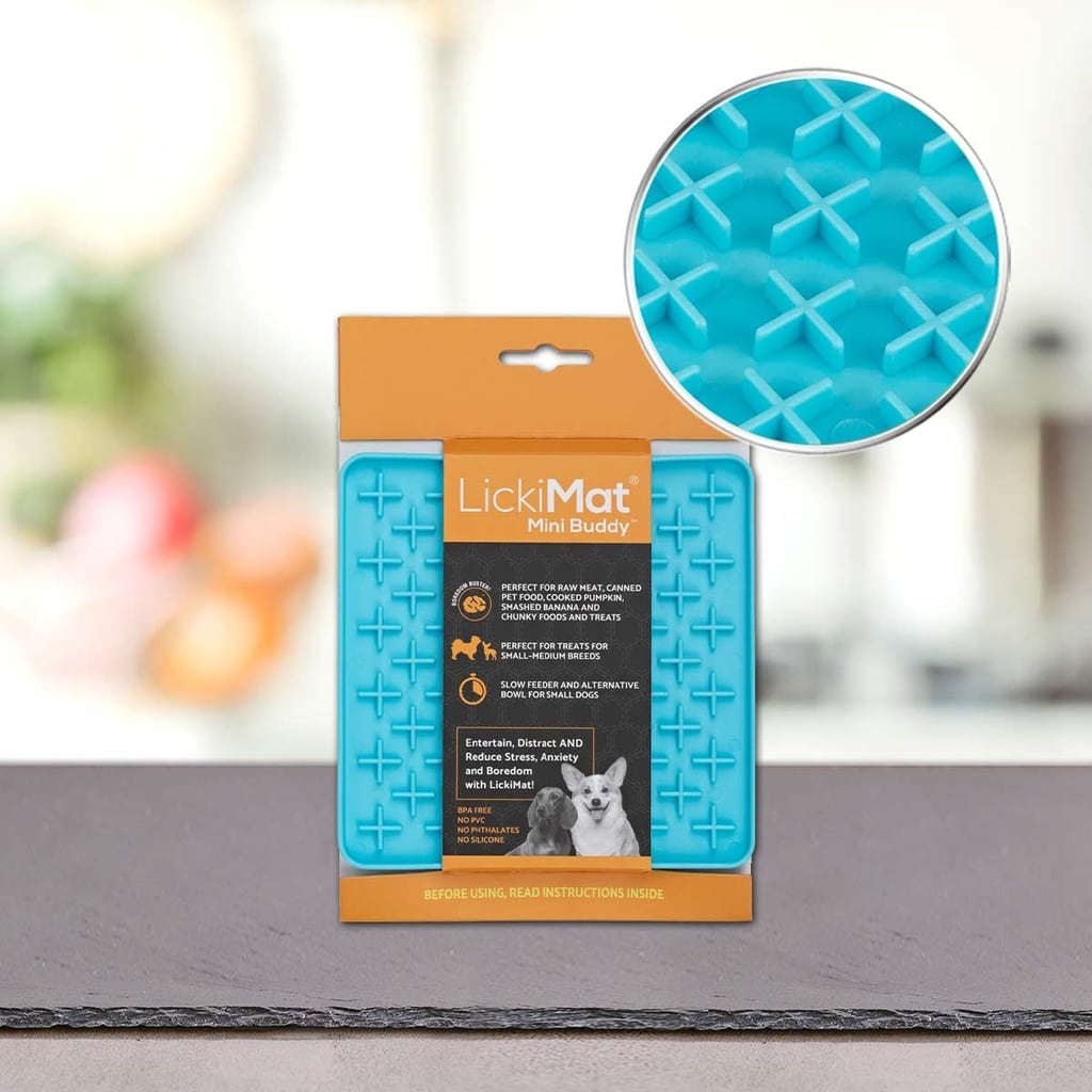 Modra Podloga LickiMat® - BUDDY by PLATINUM v oranžni embalaži stoji na mizi in prikazuje informacije o izdelku in sliko psa. Vložek poudarja njeno teksturirano površino za počasno hranjenje psa.