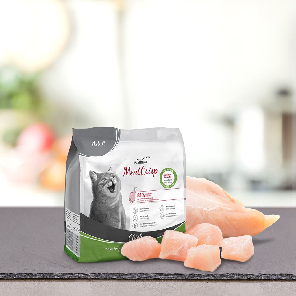 Vrečka PLATINUM MeatCrisp Adult Chicken, Suha Hrana za Mačke leži na sivi površini poleg surovih piščančjih prsi in koščkov. Na embalaži je upodobljena srečna mačka in poudarjen postopek Freshmeatdryer za odrasle mačke z okusom piščanca.