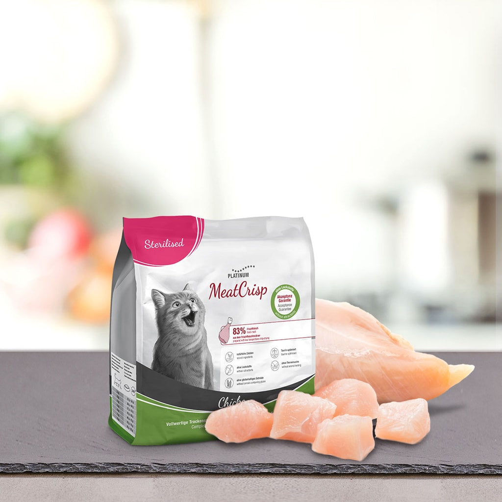 Vrečka PLATINUM MeatCrisp Sterilised Chicken Suha Hrana za Mačke s sivo mačko na sprednji strani stoji na pultu poleg kosov piščančjih prsi sveže meso na zamegljenem kuhinjskem ozadju.
