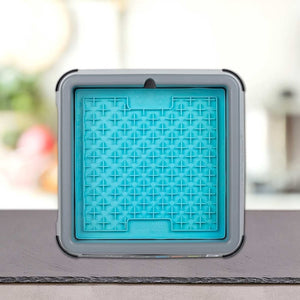 Modra kvadratna silikonska podloga za lizanje z dvignjenim križnim vzorcem PLATINUM je v sivem okvirju LickiMat® - BUDDY KEEPER na pultu z mehko zamegljenim kuhinjskim ozadjem.