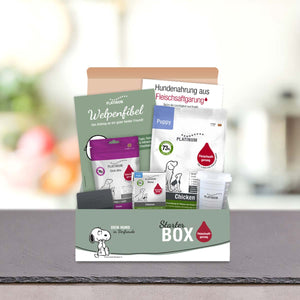 Športni paket Puppy by PLATINUM vključuje različne izdelke, brošure in vzorce za mladiče. Na škatli so ilustracije psov in nemško besedilo, prikazane na površini z zamegljenim ozadjem.