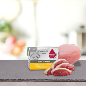 Paket PLATINUM MENU Mini mokra hrana & DoggyCoach - Nagrada za tvojega psa je prikazan na površini iz skrilavca z rezinami surove race in purana na zamegljenem kuhinjskem ozadju.