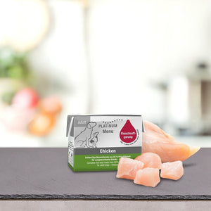 Škatla PLATINUM MENU Mini mokra hrana & DoggyCoach - Nagrada za tvojega psa stoji na pultu poleg surovega piščanca, v ozadju pa je zamegljena kuhinja. Na embalaži je ilustracija psa in zeleno-bela oblika.